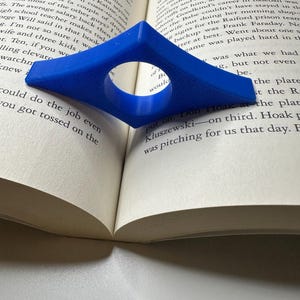 May include: A blue diamond-shaped book page holder with a circular opening, resting on the open pages of a book. The book's pages are off-white, and the text is visible. The page holder is made of plastic and is designed to keep a book open.