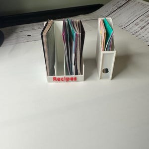 May include: Two white plastic recipe card holders. One has three compartments and the word "Recipes" in red. The other has one compartment. Both are filled with colorful recipe cards. The holders are on a white surface.
