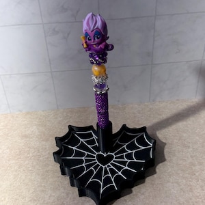 May include: A purple pen featuring a stylized character topper with white hair, accented by purple, yellow, and silver beads. The pen stands upright in a black heart-shaped base decorated with a white spiderweb pattern and a central heart outline.
