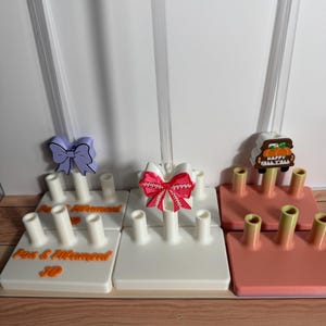 Op de afbeelding: Een verzameling strikhouders in verschillende kleuren. De houders zijn wit, koraal en oranje, elk met meerdere pinnen om strikken vast te houden. De houders hebben decoratieve strikken in paars, rood en een herfstthema. De tekst "Fox & Mermaid 3D" staat op één houder.