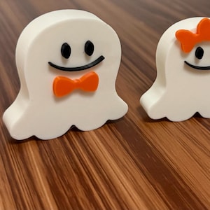 May include: Two white ghost-shaped figurines with black eyes and smiles. One has an orange bow tie, and the other has an orange bow. The figures are on a wooden surface, perfect for Halloween decorations or themed gifts.