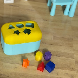 May include: A two-tone shape sorter toy with a yellow top and a light blue base. The top has cut-out shapes. Included are orange star, yellow cylinder, purple cross, red pyramid, and blue cube shapes. The toy is on a wooden surface.