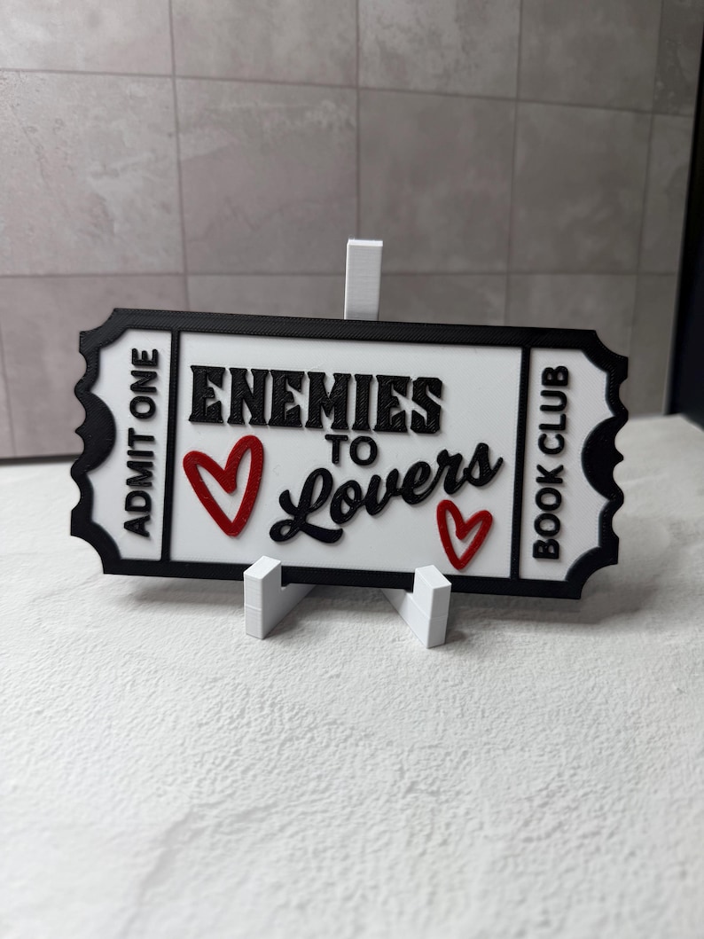 May include: A white and black sign with the text "Enemies to Lovers Book Club" and two red hearts. The sign is in the shape of a ticket and is propped up by a white stand.