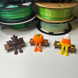 May include: Three whimsical 3D-printed figures: a brown maple leaf, an orange pumpkin, and a candy corn, each seated on a miniature wooden bench. Spools of colorful 3D printing filament are stacked in the background.
