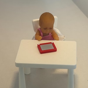 May include: A miniature doll of a baby wearing a pink outfit sits at a small white table with a red drawing toy. The baby doll is seated in a white chair. The scene is set against a white background.