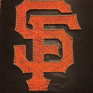 Puede incluir: Arte de hilo naranja de las letras "SF" sobre un fondo negro. Las letras están delineadas con pequeños clavos dorados, y el hilo naranja está tejido entre ellas. La obra de arte es probablemente una pieza decorativa o un artículo deportivo.