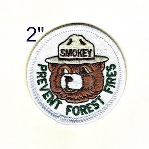 May include: A 2-inch round white embroidered patch featuring Smokey Bear wearing a ranger hat with the text "Smokey" on it. The bear is surrounded by a green circle with the text "Prevent Forest Fires".