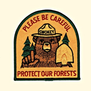 Op de afbeelding: Een geborduurde patch in groen, geel en bruin met Smokey Bear die een schop vasthoudt. De tekst luidt "Please Be Careful Protect Our Forests" en "Smokey" op de hoed van de beer.