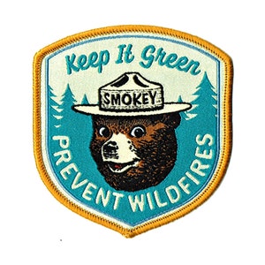 Puede incluir: Un parche bordado amarillo y azul con un oso de dibujos animados que lleva un sombrero de guardabosques. El texto en el parche dice "Keep It Green Prevent Wildfires" y "Smokey".