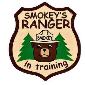 以下が含まれることがあります： 盾型のパッチ。レンジャーハットを被った茶色のクマ、スモーキーベアが描かれています。パッチには茶色の文字で「Smokey's Ranger in Training」と書かれています。緑の松の木がスモーキーの顔を挟み、ベージュの背景に配置されています。