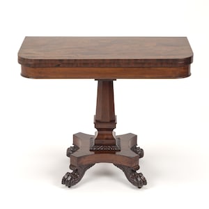 May include: A dark brown wooden pedestal table with a rectangular top featuring rounded corners. The table is supported by a central column that rests on a four-legged base, with each leg ending in a carved claw foot design.