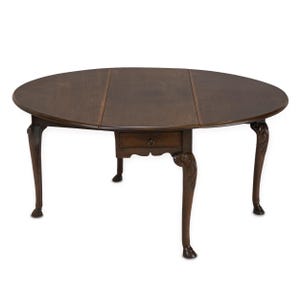 May include: A dark brown, oval-shaped antique table with a drawer. The table has a drop-leaf design and stands on four carved legs. The legs have a claw-and-ball foot design. The table is approximately 48 inches in diameter when fully extended.