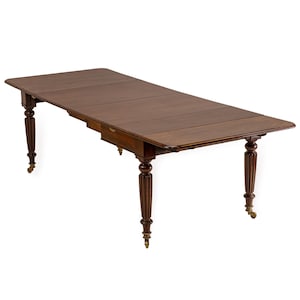 Op de afbeelding: Een donkerbruine, rechthoekige eettafel met een uitschuifbare lade. De tafel heeft vier sierlijke, gedraaide poten met messing wieltjes. De tafel is ongeveer 183 cm lang wanneer volledig uitgeschoven.