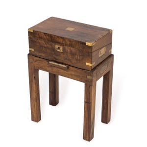 Op de afbeelding: Een donkerhouten kist met messing details, geplaatst op een bijpassende houten standaard. De kist heeft een sleutelgat en messing hoekbeschermers. De standaard heeft een rechthoekige bovenkant en vier poten, wat een klassieke, antieke uitstraling geeft.