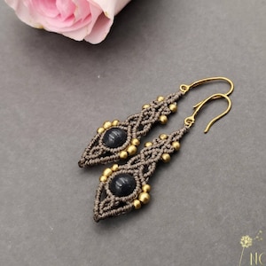 Könnte beinhalten: Graue Makrame-Ohrringe mit goldenen Perlen und dunkelblauen Edelstein-Akzenten. Die Ohrringe hängen an goldenen Haken.