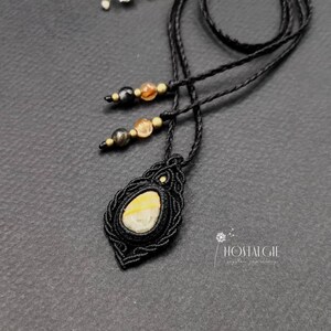 Könnte beinhalten: Eine schwarze Makramee-Kette mit einem gelben und weißen Achat-Anhänger. Die Kette hat eine schwarze Schnur und goldene Perlen. Der Anhänger ist tropfenförmig und von einem Makramee-Muster umgeben. Die Kette liegt auf einem schwarzen Hintergrund. "Nostalgie" steht in weißer Schrift unten rechts im Bild.