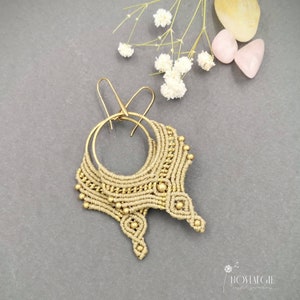 May include: A pair of gold hoop earrings with a beige macrame design and gold beads. The earrings are hanging from a gray surface.