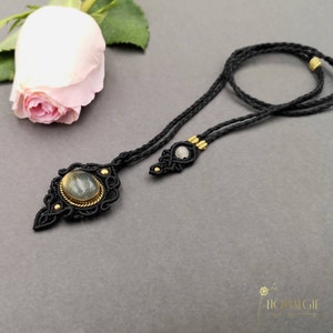 Könnte beinhalten: Eine schwarze Makramee-Kette mit einem goldfarbenen Anhänger, der mit einem grünen Edelstein besetzt ist. Der Anhänger hat die Form einer Träne mit filigranen Details. Die Kette liegt auf einer grauen Oberfläche mit einer rosa Rose im Hintergrund.