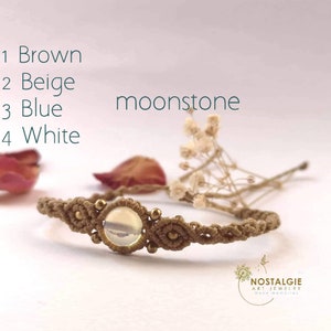 Könnte beinhalten: Ein braunes Makramee-Armband mit einem Mondstein-Perle. Das Armband ist aus einem geknüpften Band gefertigt und hat goldene Akzente. Die Mondstein-Perle ist irisierend und hat eine weiße und graue Farbe.