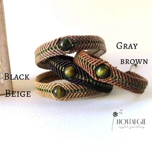 Puede incluir: Cuatro pulseras trenzadas con rayas verdes y marrones y una cuenta de ojo de tigre verde. Las pulseras están apiladas una encima de la otra en el siguiente orden: beige, negro, marrón y gris. El texto "NOSTALGIE crystal jewellery" es visible en la esquina inferior derecha de la imagen.