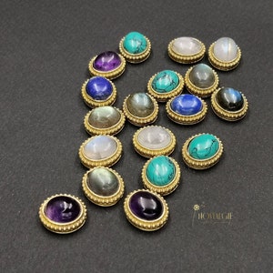 Könnte beinhalten: Sortiment ovaler Edelstein-Cabochons, gefasst in vergoldeten Zargen. Die Steine umfassen Amethyst, Türkis, Mondstein, Labradorit und Lapislazuli.