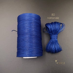 Puede incluir: Una bobina de hilo encerado azul oscuro, etiquetada "80 nationale", con una pequeña muestra de hilo a su lado.