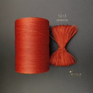 Puede incluir: Un carrete de hilo naranja y un manojo de hilo suelto se muestran sobre una superficie oscura. El carrete es cilíndrico y el hilo suelto está atado. El texto "5213 arancio" es visible.
