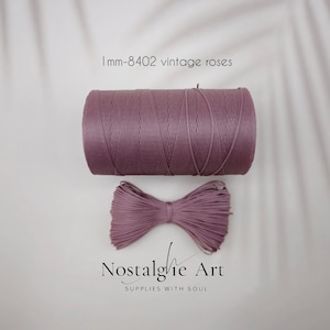 May include: A spool of mauve-colored cord and a matching bow, with the text "1mm-8402 vintage roses" and "Nostalgie Art SUPPLIES WITH SOUL". The cord is tightly wound, and the bow is neatly tied.