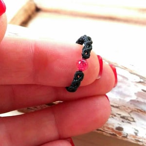 Könnte beinhalten: Ein schwarzer Schnurarmband mit zwei roten Edelsteinperlen. Der Ring befindet sich an einem Finger mit rotem Nagellack.