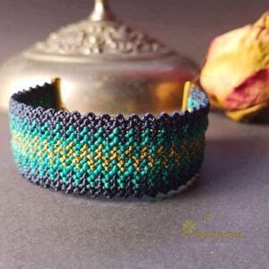 Puede incluir: Un brazalete tejido con un patrón geométrico en tonos de verde azulado, dorado y azul marino. El brazalete está hecho con una técnica de macramé y tiene un diseño simple y elegante.