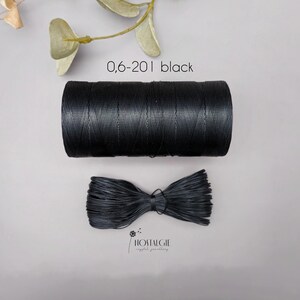 Könnte beinhalten: Eine Spule schwarzen Fadens und ein gebündeltes Bündel desselben Fadens werden auf grauem Hintergrund gezeigt. Der Text "0,6-201 black" befindet sich über der Spule. Das Logo "HOSTALGIE" befindet sich unten.