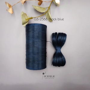 Puede incluir: Un carrete de hilo azul oscuro y un manojo del mismo hilo, atados. El texto "0,6-2568 black blue" es visible. La imagen también incluye una ramita de hojas y el logotipo "NOSTALGIE crystal jewellery".