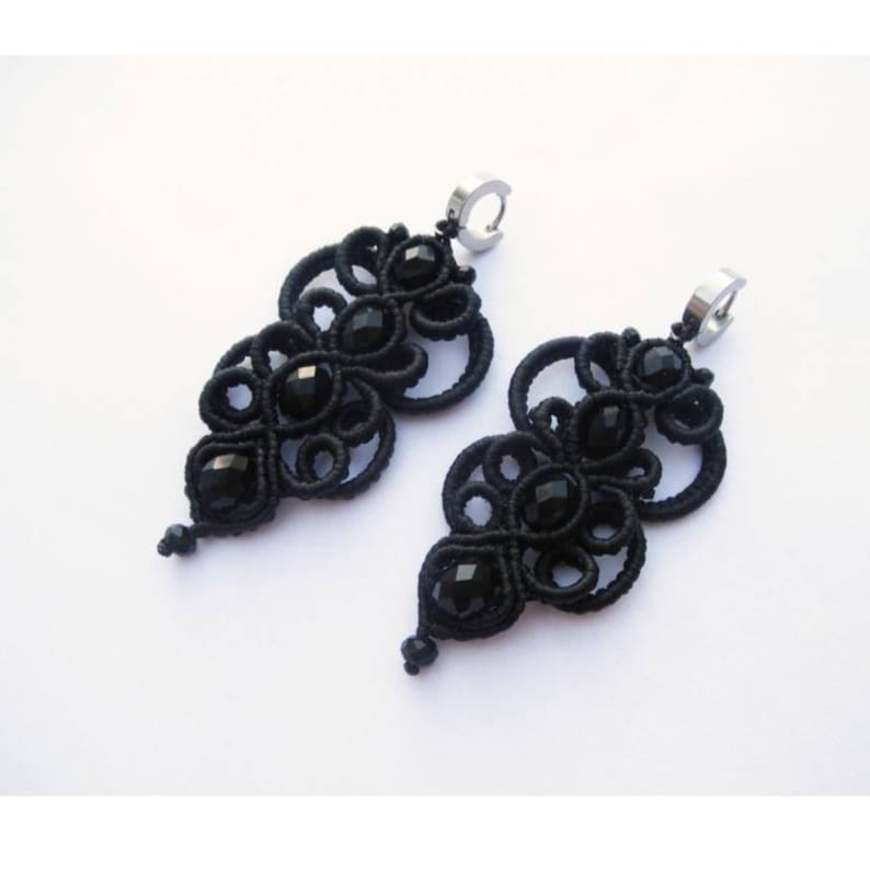 May include: A pair of black earrings with intricate knotted designs and black crystal beads. The earrings are made with black thread and have a silver hook fastening.