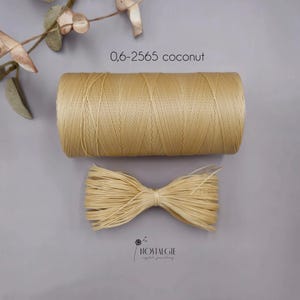 Könnte beinhalten: Eine Spule hellbraunen Fadens und ein Bündel ähnlichen Materials werden vor grauem Hintergrund gezeigt. Der Text "0,6-2565 coconut" befindet sich über der Spule. Das Logo "NOSTALGIE crystal jewellery" ist unten.