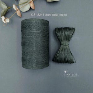 Puede incluir: Hilo verde salvia oscuro, presentado como una bobina y un haz. El texto "0,8-8241 dark sage green" es visible. El hilo se muestra sobre una superficie gris apagada, con una ramita de vegetación en el fondo.