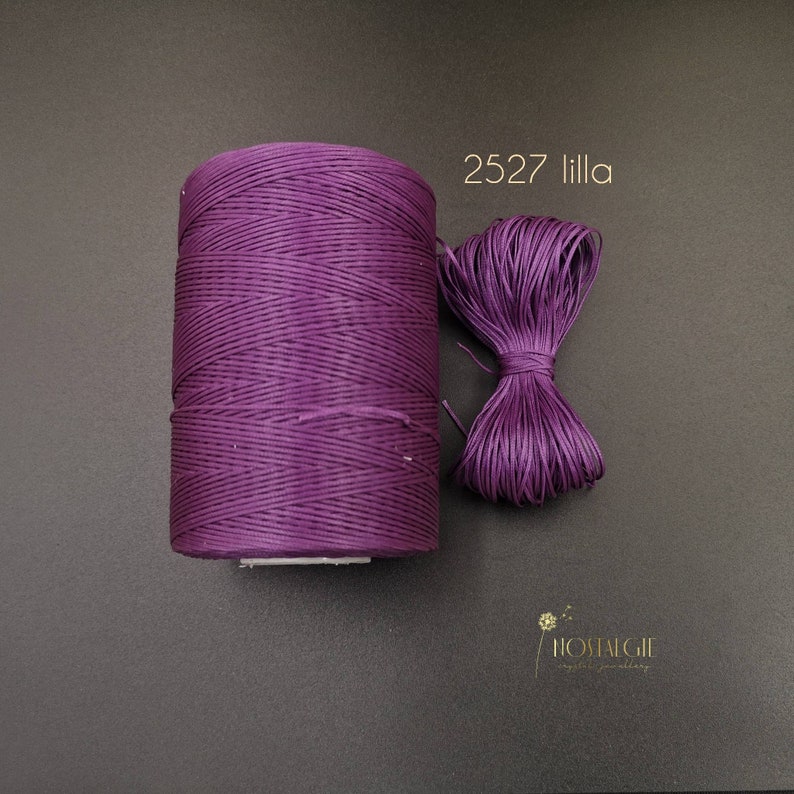 May include: A spool of purple waxed cord, labeled "2527 lilla", is shown on a dark gray surface. The cord is wound tightly and has a smooth, polished finish.