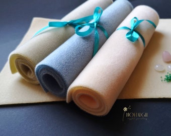 May include: Three rolls of felt in cream, blue, and beige, secured with teal ribbons. The felt rolls are displayed on a beige felt sheet, against a dark backdrop. Small crystals and the Nostalgii logo are also visible.