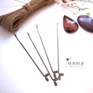 Könnte beinhalten: Drei silberne Perlennadeln mit einem gebogenen Ende zum Einfädeln von Perlen. Die Nadeln liegen auf einer weißen Oberfläche mit einer Spule braunen Kordels und zwei rosa und violetten Steinen.