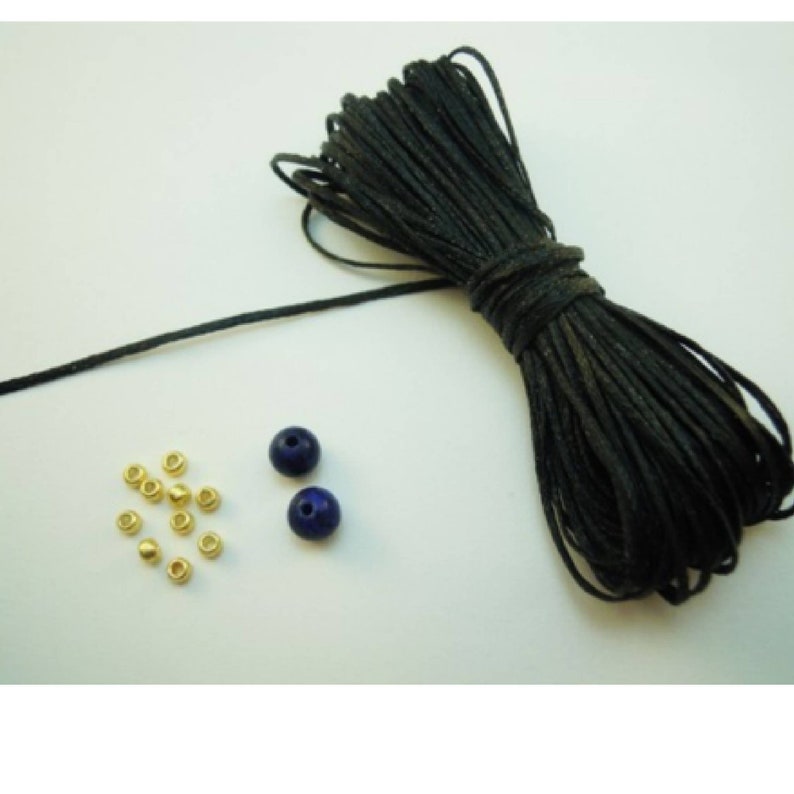 K&ouml;nnte beinhalten: Auf wei&szlig;em Hintergrund liegen eine Rolle schwarzes Kordel, zwei dunkelblaue Perlen und mehrere kleine goldfarbene Metallperlen. Die Kordel ist eng gewickelt, die Perlen sind glatt und rund. Geeignet f&uuml;r Schmuckherstellung.