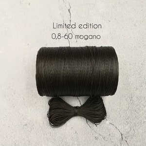 Könnte beinhalten: Eine Spule mit dunkelbraunem gewachstem Garn, beschriftet "Limited edition 0,8-60 mogano". Das Garn ist um einen Pappzylinder gewickelt und im Vordergrund befindet sich eine kleine Schleife aus dem Garn.