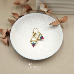 Könnte beinhalten: Goldfarbene Ohrringe mit geometrischem Design. Jeder Ohrring hat eine dreieckige Form, die mit kleinen, farbigen Perlen in Weiß, Türkis und Rosa gefüllt ist. Die Ohrringe werden auf einem weißen Teller mit einem dezenten Muster präsentiert.