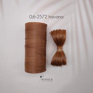 Puede incluir: Un carrete de cordón marrón y un manojo de cordón cortado se muestran sobre un fondo blanco. El texto "0,6-2572 havana" está en la parte superior y el logotipo de Nostalgie en la parte inferior. El cordón probablemente se utiliza para manualidades o joyería.