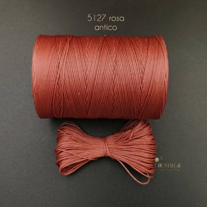 May include: A spool of red waxed cord, labeled "5127 rosa antico", with a small bow of the same cord in front of it.