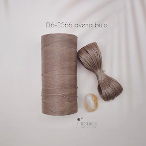 Puede incluir: Un carrete de hilo color beige y un manojo de hilo suelto se muestran junto a una pequeña piedra ovalada amarilla y blanca. El texto "0,6-2566 avena buio" está en la parte superior. El logotipo de Nostalgie crystal jewellery está en la parte inferior.