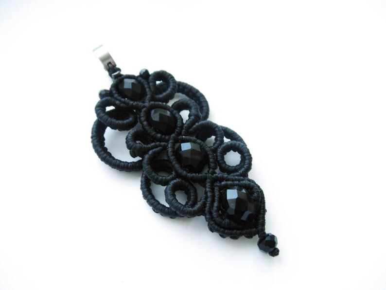 May include: Black pendant necklace with a delicate, intricate design made of black cord and black beads. The pendant is shaped like a teardrop and features a repeating pattern of circles and loops.