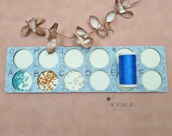 May include: A grey felt bead organiser with 12 compartments labelled A to L. The compartments contain various colours of beads, and a spool of blue thread is in compartment K. The organiser has the text "NOSTALGIE" printed on the bottom.
