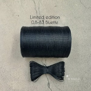 Könnte beinhalten: Eine Spule schwarzen gewachsten Fadens mit der Aufschrift "Limited edition 0,8-83 Bluette". Der Faden ist zu einer ordentlichen Spule gewickelt und auf einer hellgrauen Oberfläche abgelegt. Eine kleine Schleife aus dem gleichen Faden ist ebenfalls abgebildet.