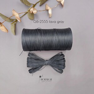 May include: A dark grey thread spool and a tied bundle of the same thread are displayed. The thread is labelled "0,6-2555 lava gray". The thread is a muted charcoal colour. The word "NOSTALGIE" is printed at the bottom.