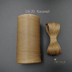 Puede incluir: Una bobina de hilo encerado Karamell 0,8-20, un tipo de cordón utilizado para la elaboración de cuero, se muestra sobre una superficie gris. El hilo es marrón y está enrollado alrededor de una bobina blanca. La bobina está etiquetada "0,8-20 Karamell 5".