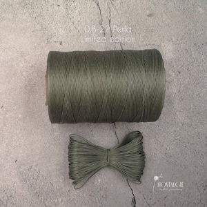May include: A spool of dark green waxed cord, labeled "0.8-22 Perla Limited edition", with a bow of the same cord in front of it.